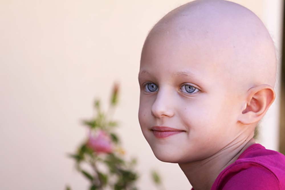 A child with a pink shirt and a bald head from chemo looks off to the left with a slight smile on their face. In the background a branch of a flowering bush is visible.