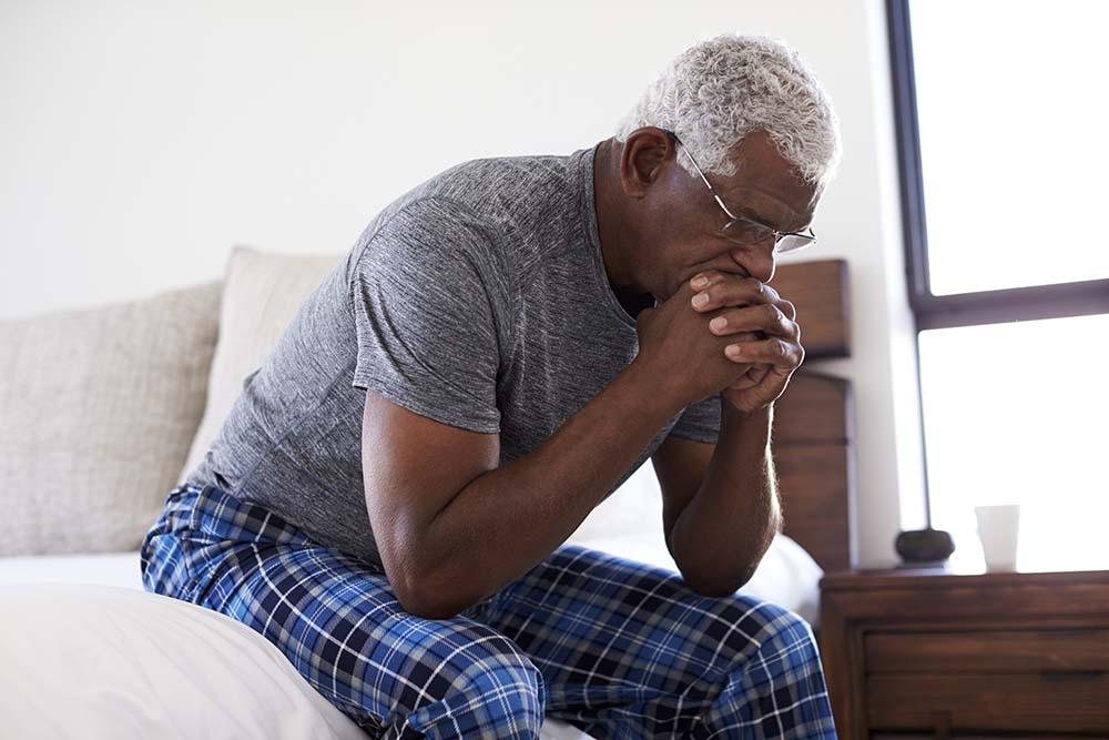 een oude zwarte man met kort wit haar in pyjama zit op de rand van het bed, vermoeid, met zijn gezicht in zijn handen.